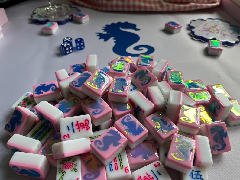 Puede incluir: Un conjunto de fichas de Mahjong rosas y blancas con dise&ntilde;os de caballitos de mar y caracteres chinos. Las fichas est&aacute;n dispuestas sobre una superficie blanca, con una silueta de caballito de mar azul en el fondo. Incluye dados azules y elementos decorativos.