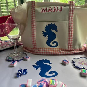 Puede incluir: Un bolso tote crema con ribete de cuadros vichy rosa y un dise&ntilde;o de caballito de mar azul. La bolsa tiene las letras "MAHJ" en la parte delantera. Tambi&eacute;n se muestran fichas de Mahjong, dados, posavasos y una copa de vino, todo sobre una superficie blanca.
