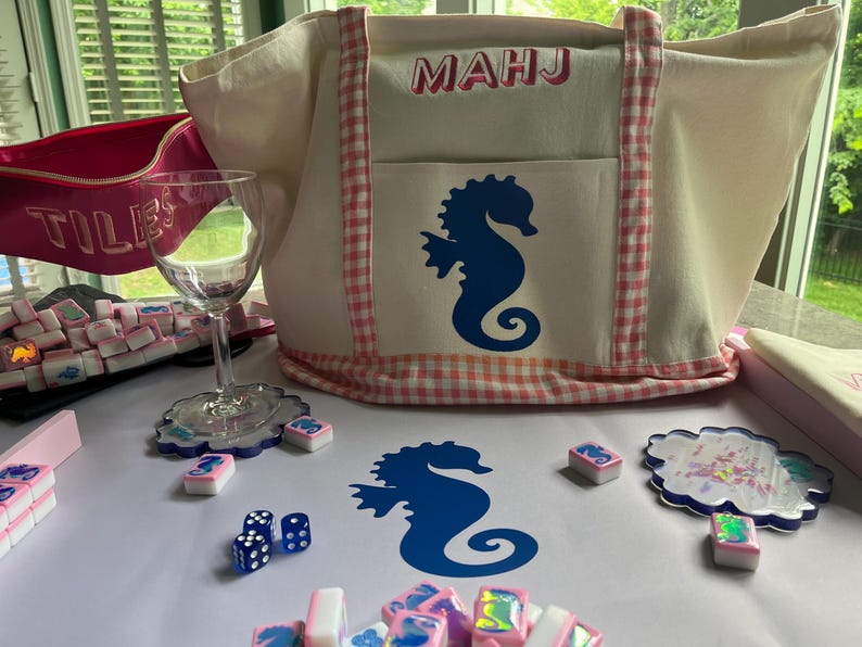 Puede incluir: Un bolso tote crema con ribete de cuadros vichy rosa y un dise&ntilde;o de caballito de mar azul. La bolsa tiene las letras "MAHJ" en la parte delantera. Tambi&eacute;n se muestran fichas de Mahjong, dados, posavasos y una copa de vino, todo sobre una superficie blanca.
