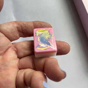 Puede incluir: Una baldosa rectangular rosa y blanca con un dise&ntilde;o abstracto colorido. La baldosa se sostiene en una mano, con otras baldosas similares visibles en el fondo. El dise&ntilde;o presenta colores azul, amarillo y blanco.