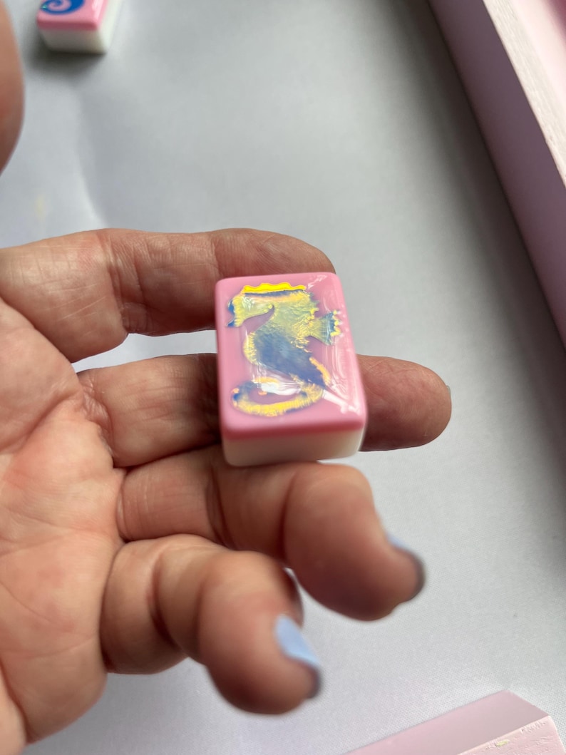 Puede incluir: Una baldosa rectangular rosa y blanca con un dise&ntilde;o abstracto colorido. La baldosa se sostiene en una mano, con otras baldosas similares visibles en el fondo. El dise&ntilde;o presenta colores azul, amarillo y blanco.