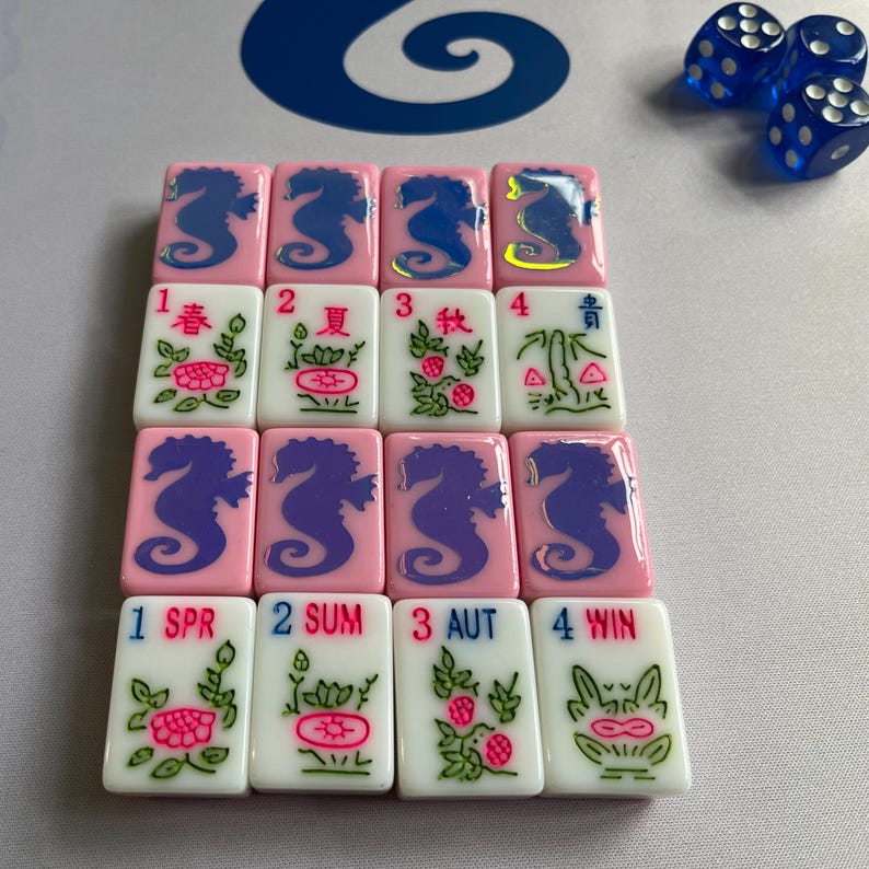 Puede incluir: Un juego de fichas de Mahjong rosas y blancas con dise&ntilde;os de caballitos de mar y flores. Las fichas presentan n&uacute;meros y s&iacute;mbolos estacionales, con dados azules en el fondo. Las piezas del juego est&aacute;n dispuestas sobre una superficie clara.