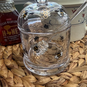May include: A clear glass honey jar with a lid and a honey dipper. The jar is decorated with bee and honeycomb designs. A bottle of Texas Wildflower honey is visible to the left.