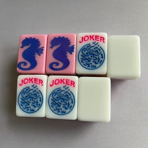 Puede incluir: Un conjunto de fichas de Mahjong. Dos fichas rosas presentan dise&ntilde;os de caballitos de mar azules. Tres fichas blancas tienen la palabra "JOKER" en rojo y dise&ntilde;os circulares azules. Dos fichas blancas en blanco completan el conjunto.