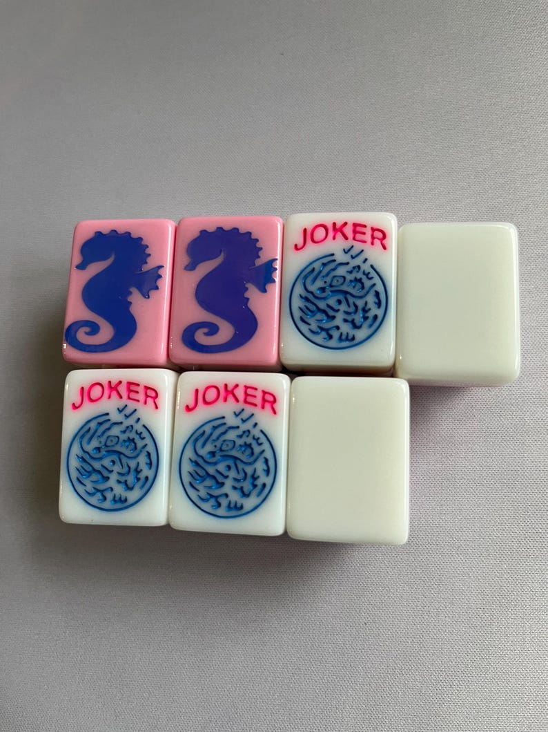 Puede incluir: Un conjunto de fichas de Mahjong. Dos fichas rosas presentan dise&ntilde;os de caballitos de mar azules. Tres fichas blancas tienen la palabra "JOKER" en rojo y dise&ntilde;os circulares azules. Dos fichas blancas en blanco completan el conjunto.