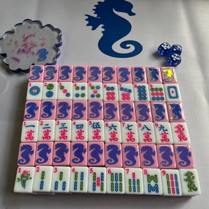 Puede incluir: Un juego de Mahjong completo con fichas rosas con dise&ntilde;os de caballitos de mar y caracteres chinos. Incluye dados azules y un posavasos decorativo con detalles rosas y morados. El juego est&aacute; dispuesto sobre una superficie blanca.