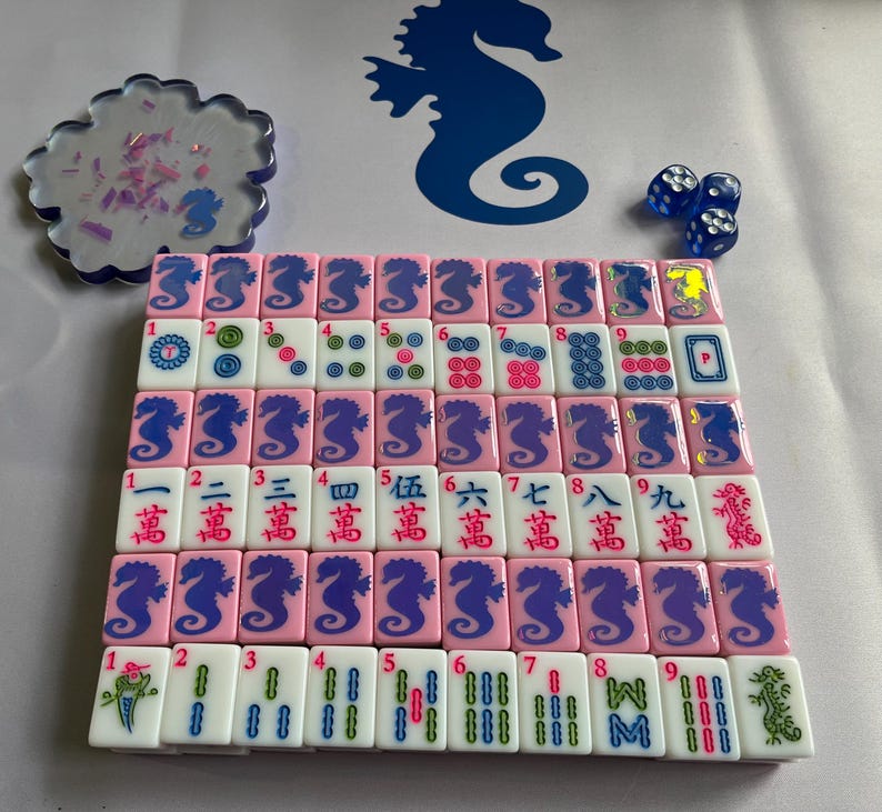 Puede incluir: Un juego de Mahjong completo con fichas rosas con dise&ntilde;os de caballitos de mar y caracteres chinos. Incluye dados azules y un posavasos decorativo con detalles rosas y morados. El juego est&aacute; dispuesto sobre una superficie blanca.