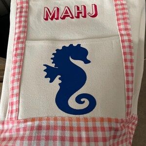 Puede incluir: Un bolso tote blanco con un dise&ntilde;o de caballito de mar. El bolso presenta una silueta de caballito de mar azul en el bolsillo delantero y las letras "MAHJ" en rojo. El bolso tiene ribetes y asas de cuadros rosas y blancos.