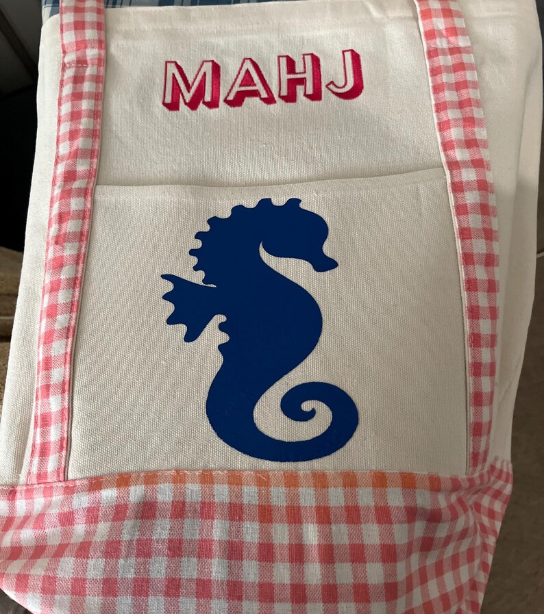 Puede incluir: Un bolso tote blanco con un dise&ntilde;o de caballito de mar. El bolso presenta una silueta de caballito de mar azul en el bolsillo delantero y las letras "MAHJ" en rojo. El bolso tiene ribetes y asas de cuadros rosas y blancos.