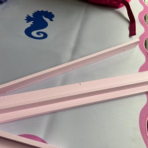 Puede incluir: Tres piezas de madera rosa claro, posiblemente para un proyecto de manualidades, est&aacute;n dispuestas sobre una superficie blanca con un borde rosa festoneado. Se ve un dise&ntilde;o de caballito de mar azul.