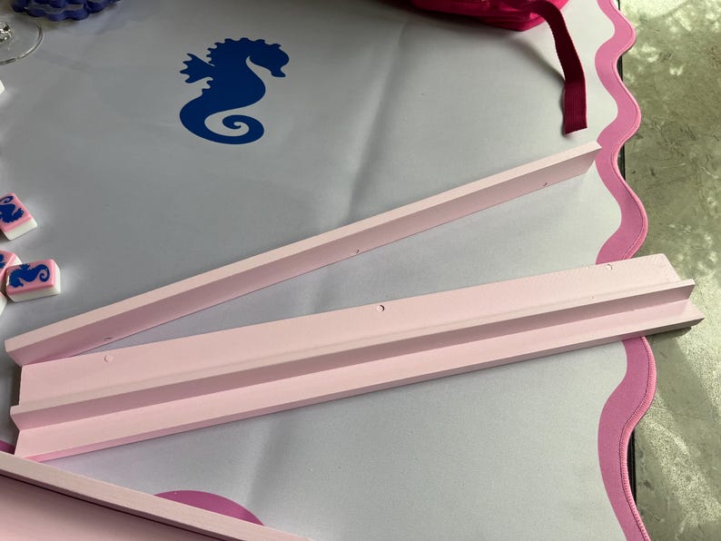 Puede incluir: Tres piezas de madera rosa claro, posiblemente para un proyecto de manualidades, est&aacute;n dispuestas sobre una superficie blanca con un borde rosa festoneado. Se ve un dise&ntilde;o de caballito de mar azul.