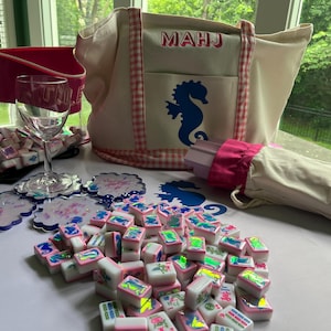Puede incluir: Un conjunto de fichas de Mahjong, posavasos, una copa de vino y un bolso de mano. El bolso de mano es beige con un ribete de cuadros rosas y un dise&ntilde;o de caballito de mar azul. Tambi&eacute;n hay una bolsa con cremallera rosa y un paraguas a juego.