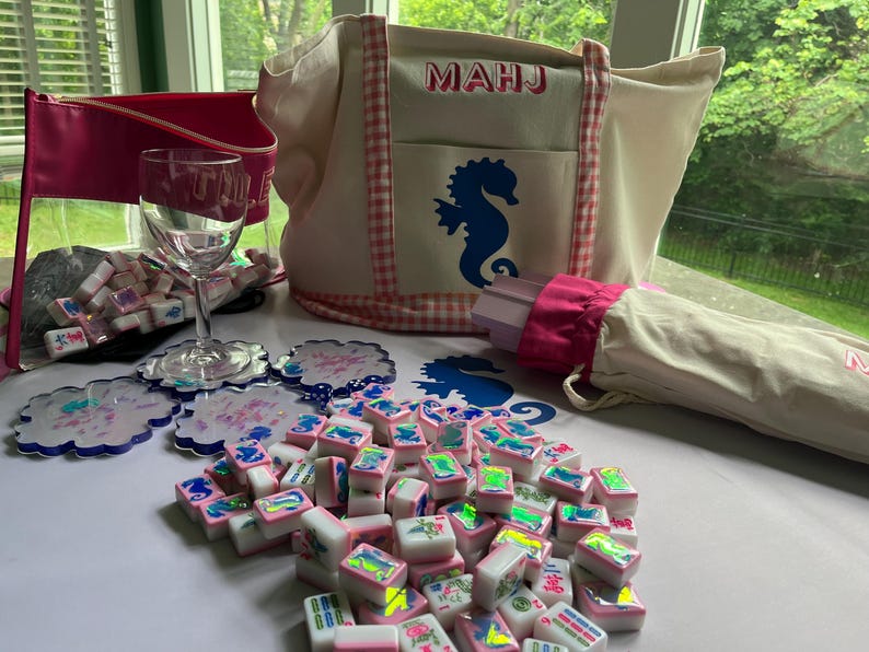 Puede incluir: Un conjunto de fichas de Mahjong, posavasos, una copa de vino y un bolso de mano. El bolso de mano es beige con un ribete de cuadros rosas y un dise&ntilde;o de caballito de mar azul. Tambi&eacute;n hay una bolsa con cremallera rosa y un paraguas a juego.