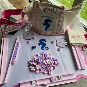 Puede incluir: Un juego de Mahjong completo con fichas rosas y blancas, un tablero de juego rosa y un bolso tote beige con "MAHJ" impreso. La bolsa presenta un dise&ntilde;o de caballito de mar. Tambi&eacute;n se incluye una bolsa con cremallera rosa y una peque&ntilde;a bolsa.