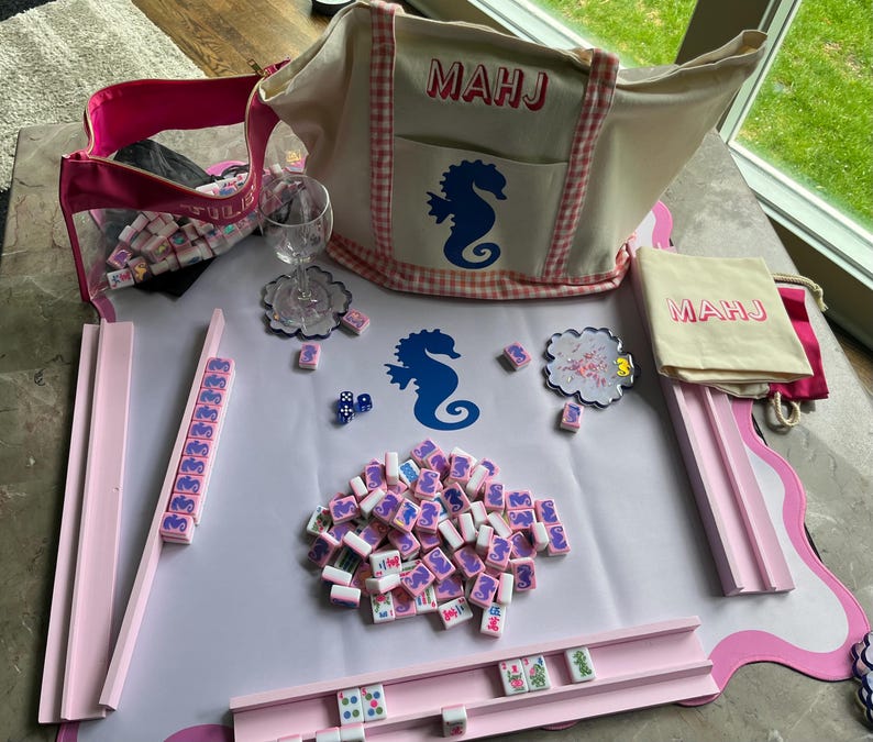 Puede incluir: Un juego de Mahjong completo con fichas rosas y blancas, un tablero de juego rosa y un bolso tote beige con "MAHJ" impreso. La bolsa presenta un dise&ntilde;o de caballito de mar. Tambi&eacute;n se incluye una bolsa con cremallera rosa y una peque&ntilde;a bolsa.