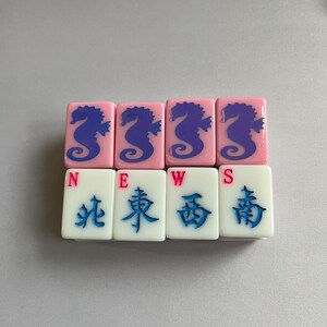 Puede incluir: Un juego de fichas de Mahjong. Cuatro fichas rosas con dise&ntilde;os de caballitos de mar azules est&aacute;n encima de cuatro fichas blancas con caracteres chinos y las letras N, E, W y S. Las fichas est&aacute;n dispuestas en dos filas.