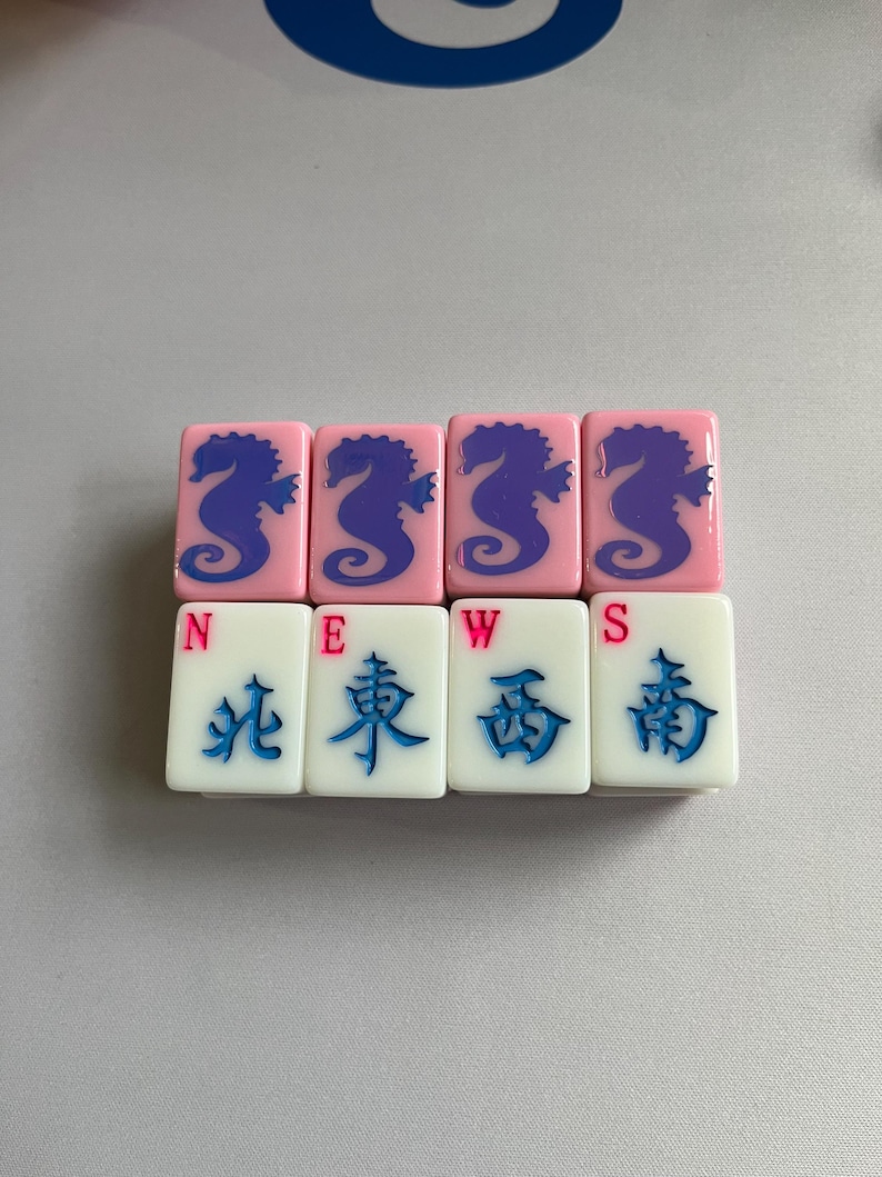 Puede incluir: Un juego de fichas de Mahjong. Cuatro fichas rosas con dise&ntilde;os de caballitos de mar azules est&aacute;n encima de cuatro fichas blancas con caracteres chinos y las letras N, E, W y S. Las fichas est&aacute;n dispuestas en dos filas.