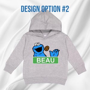 May include: A light grey hooded sweatshirt with a blue cartoon character holding a biscuit. The character has large eyes and a wide smile. The name "BEAU" is displayed on a green rectangular sign below the character. The text "DESIGN OPTION #2" is above the hood.