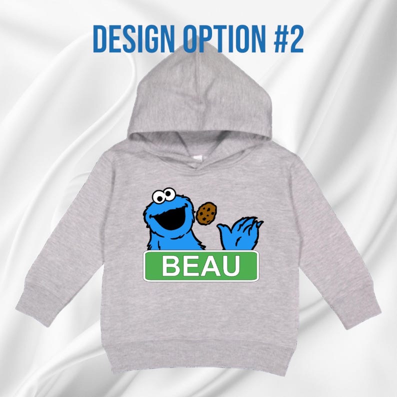 May include: A light grey hooded sweatshirt with a blue cartoon character holding a biscuit. The character has large eyes and a wide smile. The name "BEAU" is displayed on a green rectangular sign below the character. The text "DESIGN OPTION #2" is above the hood.