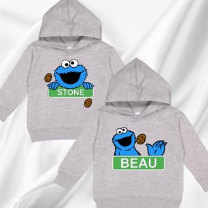 May include: Two light grey hooded sweatshirts. Each features a blue monster graphic with a cookie and a name on a green sign. One says "STONE" and the other says "BEAU".