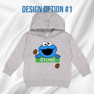 May include: A light grey hooded sweatshirt with a blue cartoon character, large mouth and googly eyes. The character holds a green sign saying "STONE" and has two cookies. The text "DESIGN OPTION #1" is above the hood.