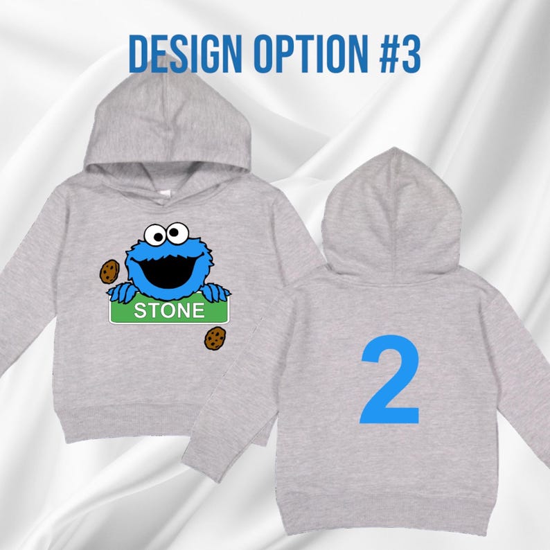 May include: A light grey hooded sweatshirt featuring a blue Cookie Monster graphic holding a green sign that reads "STONE". The back of the sweatshirt has a large blue number "2". The text "DESIGN OPTION #3" is at the top.