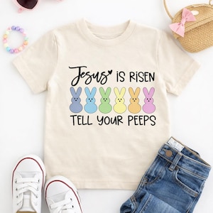 May include: A beige short-sleeved t-shirt with the text "Jesus is Risen Tell Your Peeps" and colorful bunny graphics. The image also includes a pair of white sneakers, blue jeans, pink heart-shaped sunglasses, and a small straw purse.