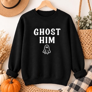May include: A black long-sleeve crewneck sweatshirt featuring the white text GHOST HIM centered on the chest, positioned above a simple white line-art graphic of a ghost. The garment has ribbed cuffs and a ribbed hemline.