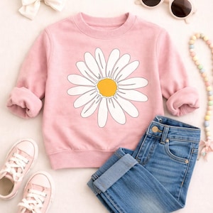 May include: A pink sweatshirt with a large white daisy graphic and a yellow center, paired with blue jeans, pink sneakers, sunglasses, a white bow, and a beaded necklace. A casual outfit for children.