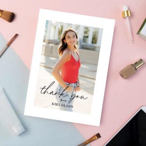 May include: A white card with a photograph of a person in a red top and jeans. The card has the words "thank you" in cursive script, with "from KIKI OLSON" below. Makeup brushes and beauty products surround the card on a pink and blue surface.