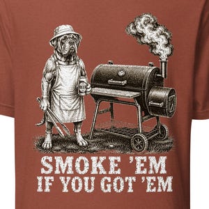 Pode incluir: Camiseta cor de tijolo com um gráfico detalhado de um buldogue com avental e chapéu, segurando uma cerveja e pinças ao lado de um fumegador. O texto "SMOKE 'EM IF YOU GOT 'EM" está impresso abaixo.
