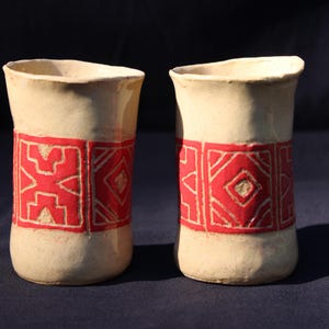 May include: Two cylindrical ceramic cups with an off-white, matte finish. Each cup features a wide, horizontal band around the center decorated with a repeating geometric pattern in a vibrant red color, contrasting against the light clay base.