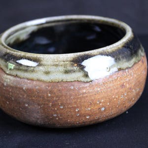 May include: A round ceramic bowl with a textured, speckled terracotta-colored base and a smooth, glossy dark brown and cream-colored glaze around the upper rim. The interior of the bowl is finished in a dark, reflective glaze.