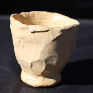 Op de afbeelding: Een kleine, met de hand gevormde keramische beker met een lichtbeige, matte afwerking. Het vat heeft een onregelmatig, gefacetteerd buitenoppervlak met een dikke, ongelijke rand en een smalle, ronde voet, wat zorgt voor een getextureerd uiterlijk.