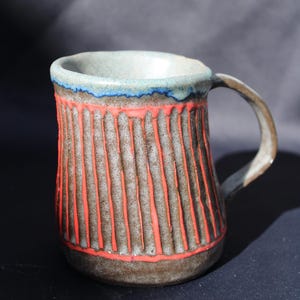 Puede incluir: Una taza de cerámica con un exterior texturizado, marrón y acanalado que presenta líneas verticales de color naranja brillante. El borde está esmaltado en azul claro con un acento azul oscuro. Tiene un asa curva y una abertura amplia.
