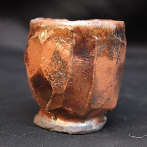 Peut inclure: Une petite tasse en céramique façonnée à la main avec une surface texturée et facettée. L'extérieur présente un émail tacheté dans des tons orange brûlé, brun profond et bronze métallique, avec un bord irrégulier et une base évasée.