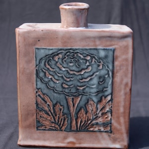 May include: A square ceramic vase with a glossy light brown finish and a central rectangular panel featuring a dark grey relief carving of a flower with textured leaves. The vase has a short, cylindrical neck at the top center.