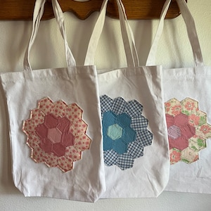 May include: Three white canvas tote bags with different patchwork flower designs. The flowers are made of fabric scraps in pink, blue, and green.