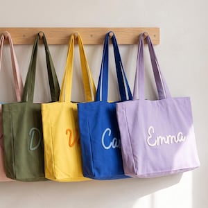 Pode incluir: Uma fila de sete sacolas de lona nas cores creme, azul claro, rosa claro, verde oliva, amarelo, azul royal e lavanda, cada uma com um nome bordado em branco em letra cursiva, penduradas em um cabideiro de madeira com sete ganchos.