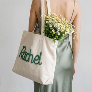 May include: Cream-colored corduroy tote bag with the name "Rachel" embroidered in green script. The bag is filled with a bouquet of white and yellow flowers. The person is wearing a sage green dress.