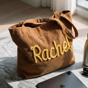 May include: A brown corduroy tote bag with the name "Rachel" embroidered in yellow. Also on the marble surface are a silver water bottle, black sunglasses, and a gray tablet.