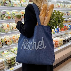 May include: A blue corduroy tote bag with the name "Rachel" embroidered in a light blue script. The bag holds two baguettes, and a bunch of fresh herbs. The person is holding a green drink and is in a grocery store.