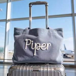 Può includere: Una borsa tote in velluto a coste blu chiaro con il nome "Piper" ricamato in bianco, appoggiata sopra una valigia argentata con ruote. La borsa ha una forma rettangolare e due manici. Lo sfondo mostra un terminal aeroportuale con un aereo.