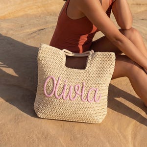 May include: A beige woven tote bag with the name "Olivia" embroidered in pink. The bag has a rectangular shape with a handle. The bag is sitting on a rock next to a person wearing a rust-coloured swimsuit.