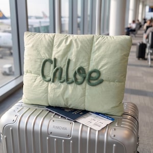 May include: A light green quilted rectangular pillow featuring the name Chloe in dark green knitted script lettering, resting on a silver metallic Rimowa suitcase with a dark blue passport and a boarding pass for a flight from LHR to JFK.