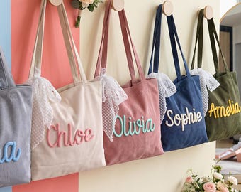 May include: Five corduroy tote bags hanging on wooden pegs against a multi-colored wall. Each bag features a white lace bow and a name embroidered in a raised script font: Luna in blue, Chloe in pink, Olivia in teal, Sophie in white, and Amelia in yellow.