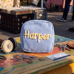 May include: A light blue corduroy square pouch featuring the name Harper embroidered in yellow yarn on the front. The pouch is positioned on a wooden surface alongside a pair of over-ear headphones, a set of metal keys, and a credit card.