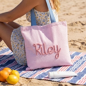 Puede incluir: Bolso tote a rayas rosas y blancas con el nombre "Riley" bordado en escritura rosa. El bolso tiene asas de mezclilla azul. La escena incluye una toalla de playa, fruta, una botella de agua y protector solar.