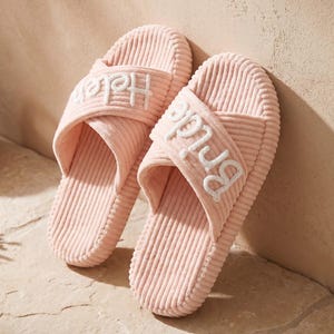 May include: A pair of light pink corduroy open-toe slippers with crisscross straps. The left slipper features the white embroidered text "Helen" and the right slipper features the white embroidered text "Bride" on the top strap.