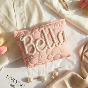 Op de afbeelding: Een roze gewatteerde cosmetische tas met de naam "Bella" in witte touwletters. De tas is omgeven door accessoires, waaronder parel haarclips, oorbellen en een zijden sjaal. Een kaart leest "FOR YOU - Bella".